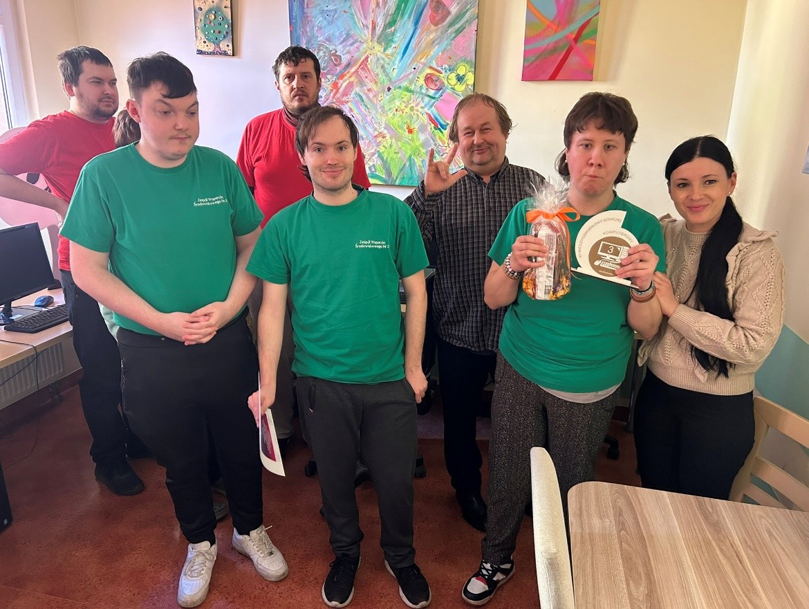 Na pierwszym planie dwóch mężczyzn i kobieta w zielonych koszulkach stoją i patrzą w obiektyw; kobieta trzyma w lewej ręce drewnianą statuetkę a w prawej upominek. W tle po lewej stronie stoją dwaj mężczyźni w czerwonych koszulkach a po prawej organizatorzy- kobieta i mężczyzna, również patrzą w obiektyw.