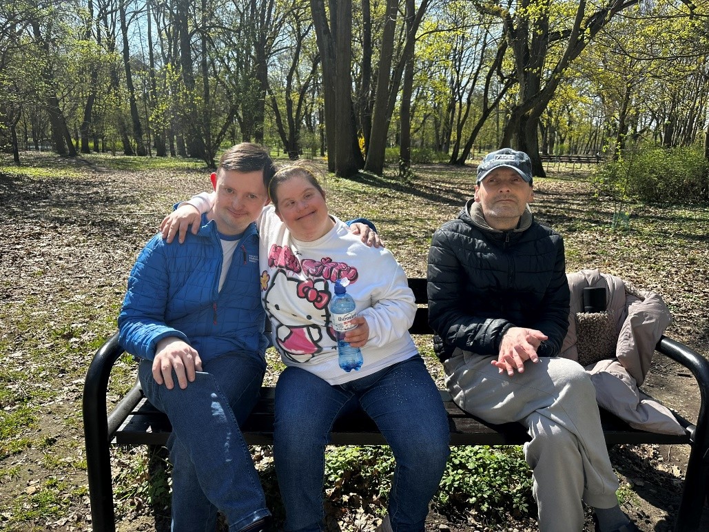 Grupa trzech uśmiechniętych osób siedzi na ławce w parku. W tle drzewa. 