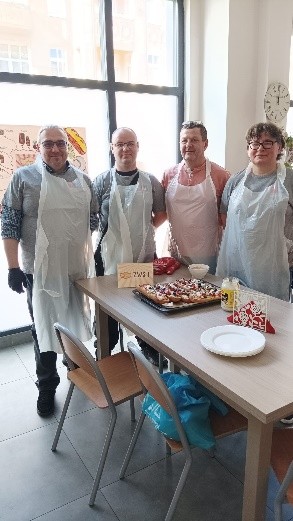 Cztery osoby stoją przy stole w jasnej kuchni. Przed nimi leży blacha z przygotowanymi zapiekankami, obok talerze, przyprawy i serwetki. W tle duże okno i zegar na ścianie.