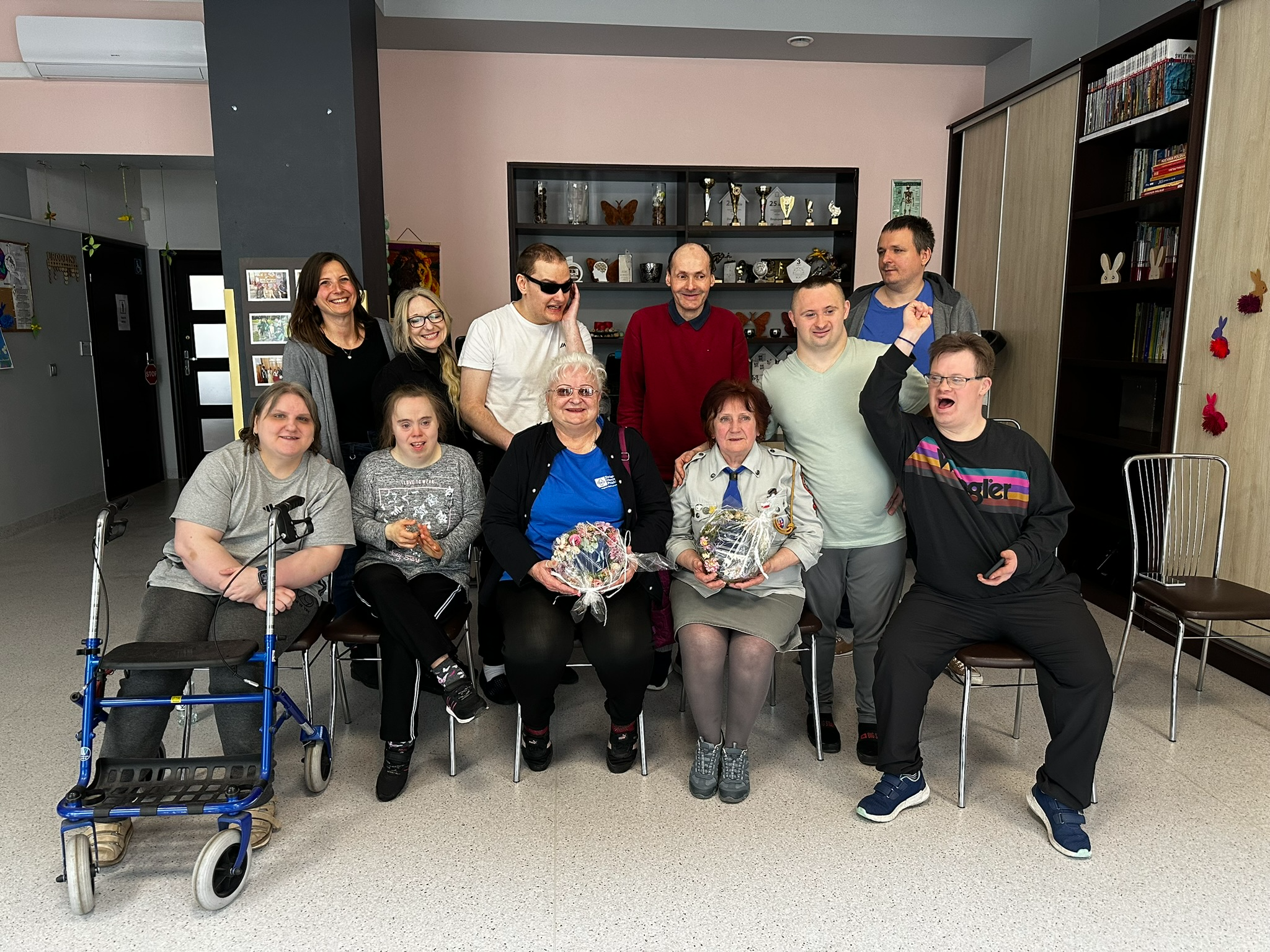 Na pierwszym planie grupa uśmiechniętych osób siedzi na krześle i patrzy prosto w obiektyw. Na drugim planie grupa uśmiechniętych osób stoi.